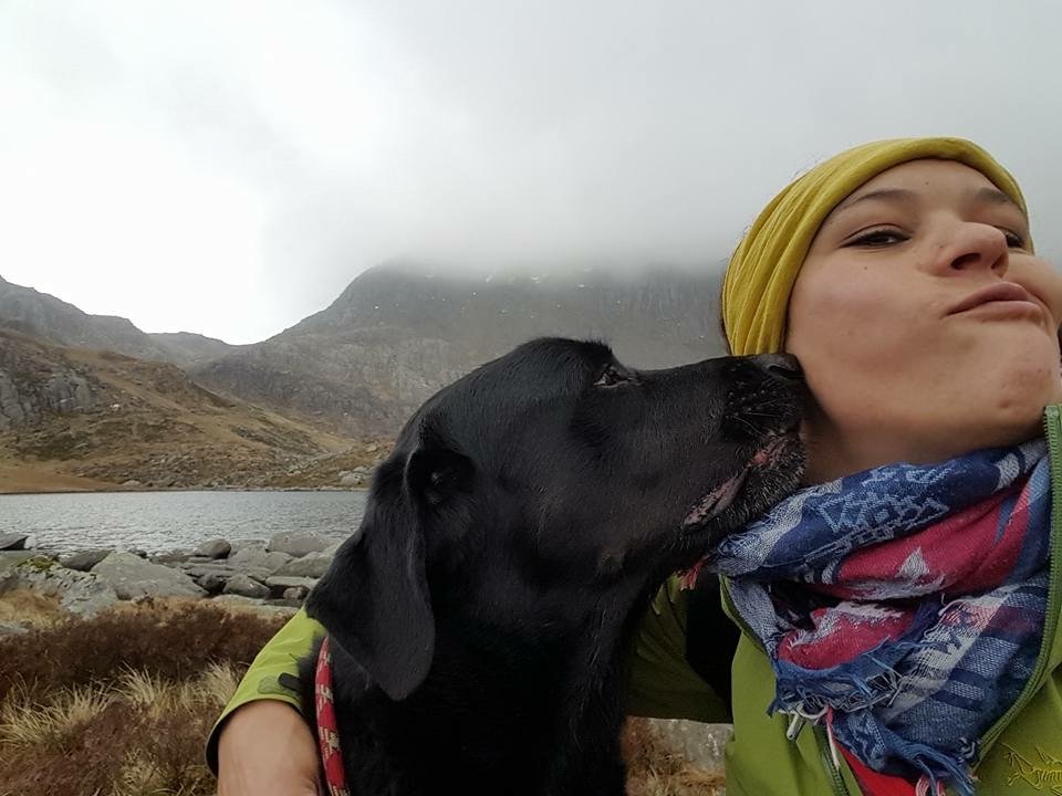 black labradorite leaning to lick a woman's face. She is wearing a yellow beanie, a blue scarf and frowning to avoid the lick on the face. mist mountains and lake in the background